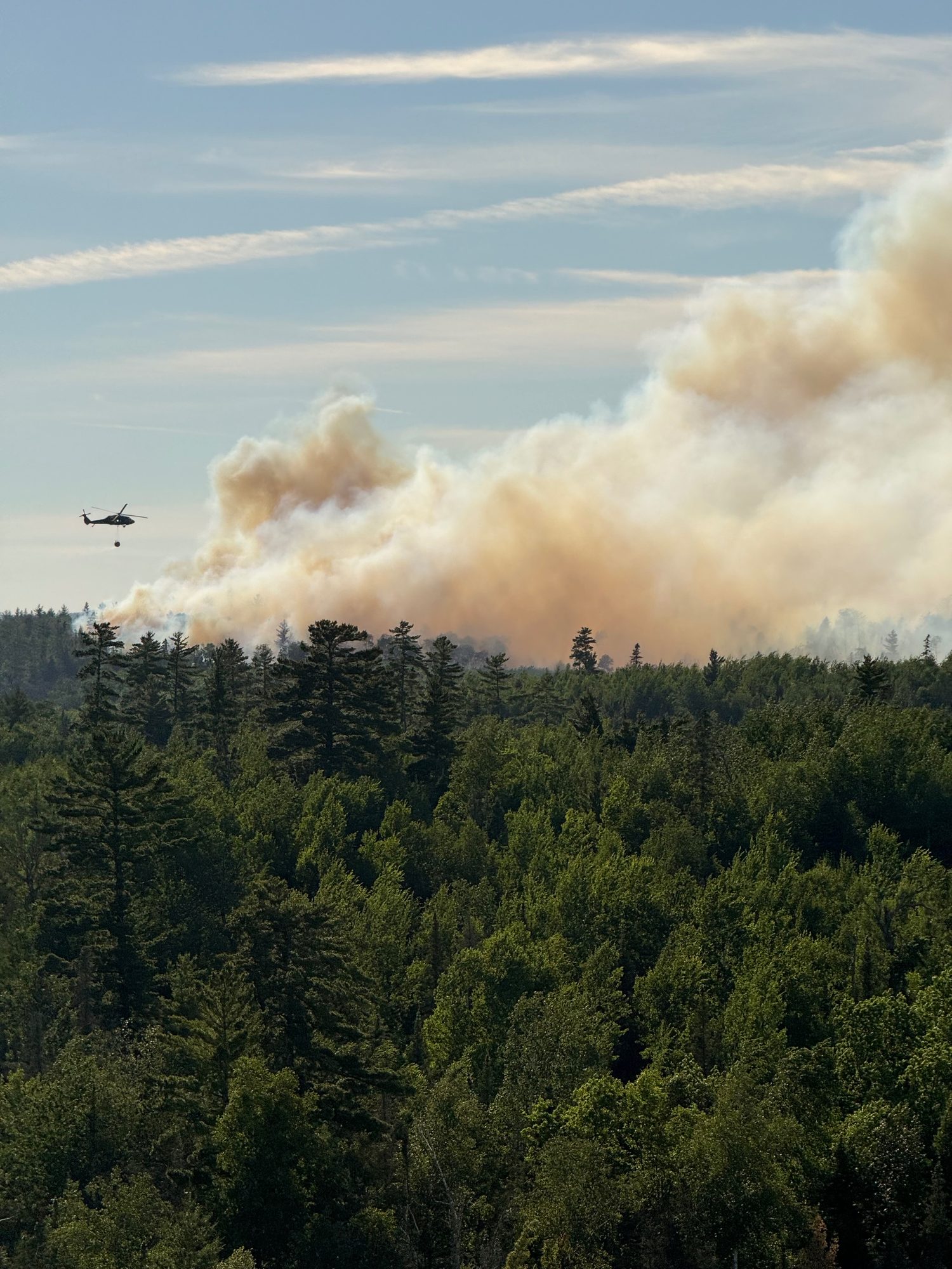 A helicopter flies toward a towering plume of tan smoke rising above a green forest canopy under a blue sky.
