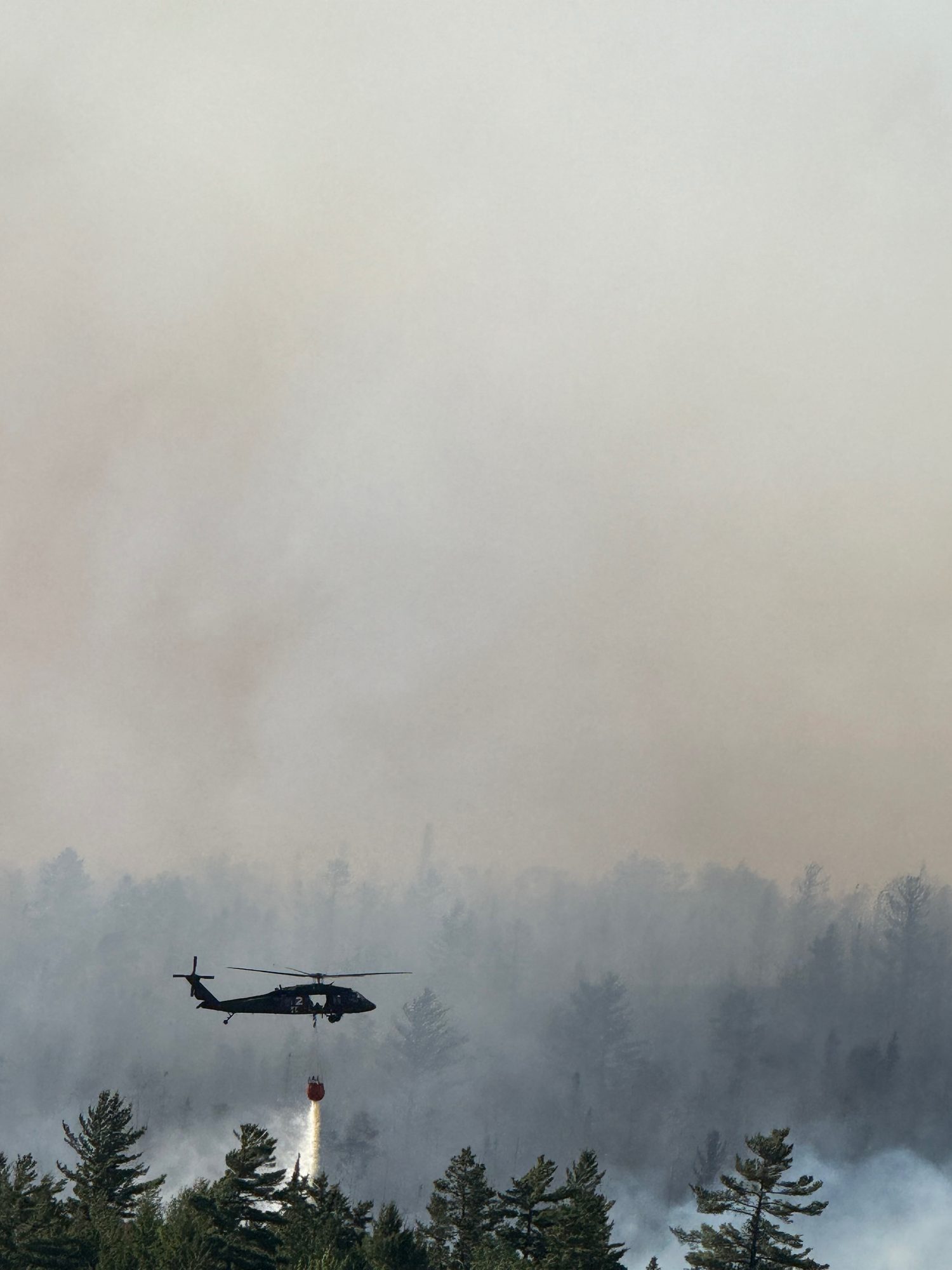 A Black Hawk helicopter releases a stream of water over a smoke-shrouded forest.